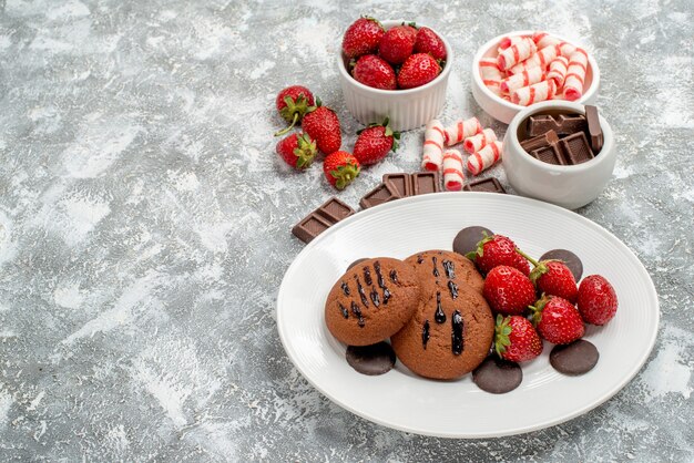 Vista inferior galletas fresas y bombones redondos en el plato blanco y cuencos de dulces fresas chocolates sobre el suelo gris-blanco