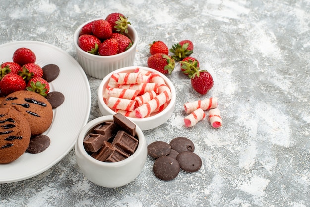 Foto gratuita vista inferior galletas fresas y chocolates redondos en el plato ovalado blanco cuencos de dulces fresas chocolates en el lado izquierdo de la mesa gris-blanca