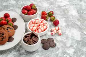 Foto gratuita vista inferior galletas fresas y chocolates redondos en el plato ovalado blanco cuencos de dulces fresas chocolates en el lado izquierdo de la mesa gris-blanca