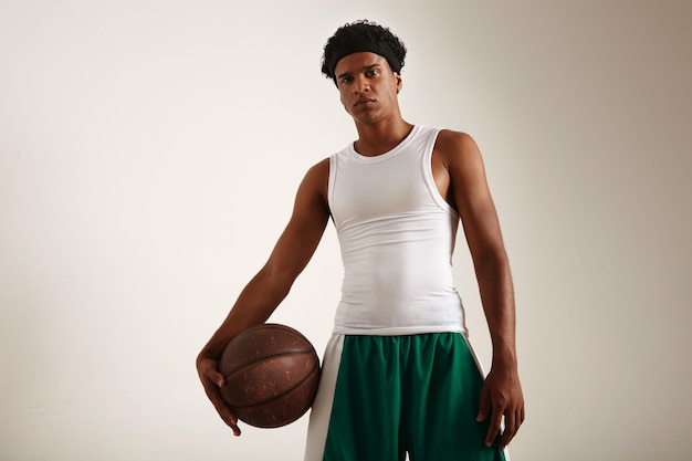 Vista inferior del jugador de baloncesto afroamericano enojado muscular tonificado en uniforme blanco y verde sosteniendo una pelota de baloncesto grunge