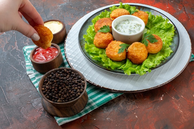 Vista inferior de nuggets de pollo, lechuga y salsa en un plato, pimienta negra en un tazón, salsas en tazones pequeños, pepita en la mano femenina en la mesa oscura.