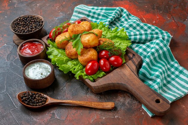 Vista inferior nuggets de pollo lechuga tomates cherry en tablero de madera pimienta negra en tazón salsas en pequeños tazones de madera cuchara de madera sobre mesa oscura