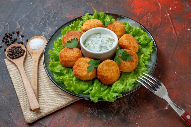 Vista inferior de nuggets de pollo, tenedor de lechuga en un plato, sal y pimienta negra en cucharas de madera en la mesa oscura.