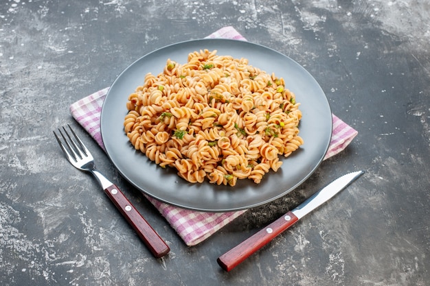 Vista inferior de la pasta rotini en un plato redondo en la servilleta a cuadros rosa blanco tenedor y cuchillo en la mesa gris