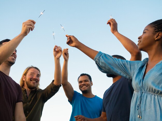 Vista inferior de personas felices de pie con luces de bengala. Sonrientes amigos pasar tiempo juntos al aire libre. Concepto de celebración