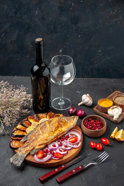 Vista inferior de pescado frito berenjenas fritas cebolla cortada en tabla de servir de madera botella de vino y tenedor y cuchillo de vidrio especias de ajo sobre fondo oscuro