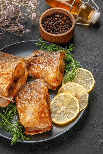 Vista inferior de pescado frito con rodajas de limón en un plato de pimienta negra en un tazón de madera botella de aceite sobre fondo oscuro