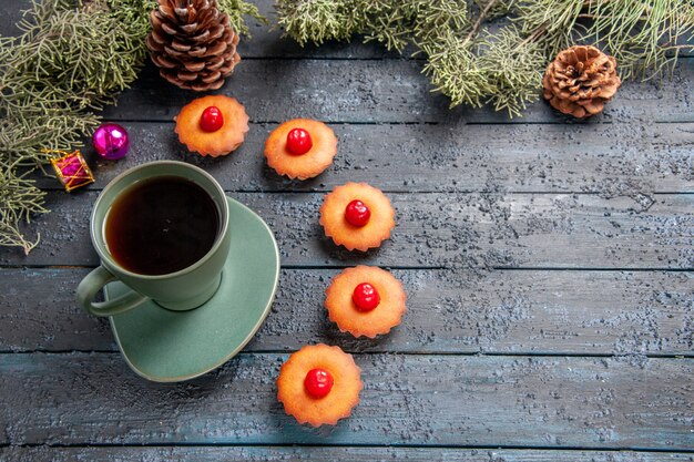 Vista inferior redondeada cereza cupcakes ramas de abeto juguetes de navidad conos y una taza de té sobre fondo de madera oscura.