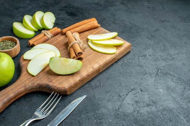 Vista inferior de rodajas de manzana y palitos de canela en una tabla de cortar, cuchillo y tenedor, polvo de menta seca en un tazón pequeño sobre una mesa oscura con espacio libre