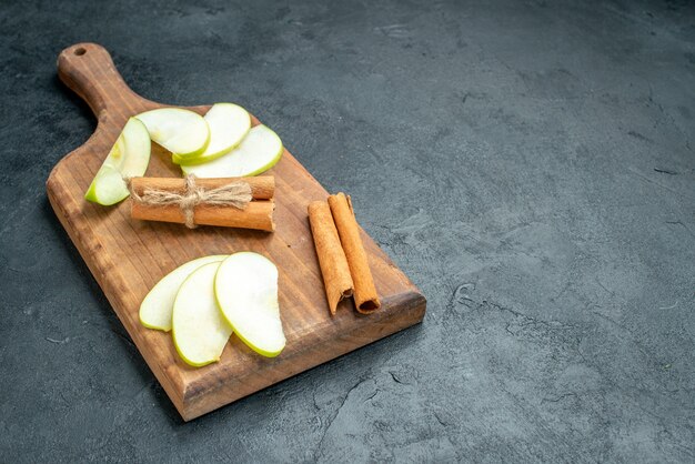 Vista inferior de rodajas de manzana y palitos de canela en una tabla de cortar en una mesa oscura con espacio libre