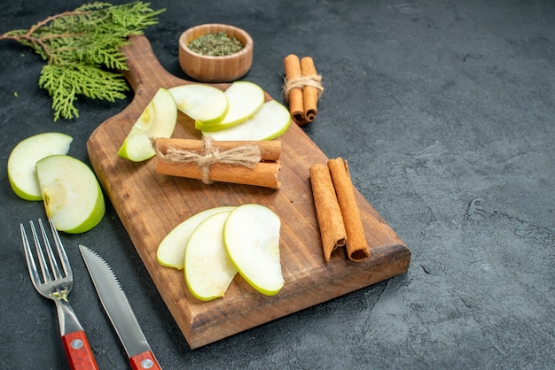 Vista inferior de rodajas de manzana y palitos de canela en un tablero de madera, cuchillo y tenedor, polvo de menta seca en un tazón pequeño sobre una mesa oscura con espacio libre