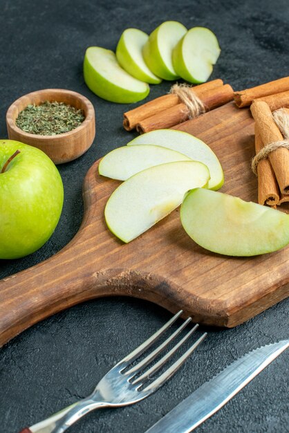 Vista inferior de rodajas de manzana y ramas de canela en la tabla de cortar, cuchillo y tenedor, polvo de menta seca en un tazón pequeño sobre una mesa oscura