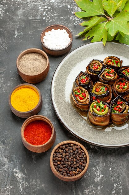 Vista inferior rollos de berenjena rellena en plato ovalado blanco diferentes especias en tazones pequeños sobre fondo gris