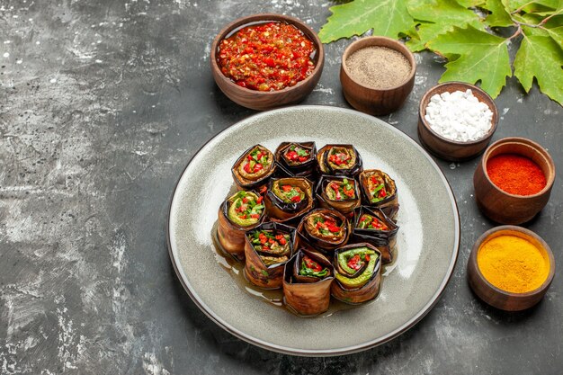 Vista inferior rollos de berenjena rellena en plato ovalado blanco especias en tazones pequeños sal pimienta pimiento rojo cúrcuma adjika sobre fondo gris lugar libre