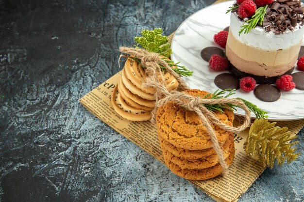 Vista inferior de tarta de queso con chocolate y frambuesas en plato ovalado blanco galletas atadas en adornos navideños de periódico sobre fondo gris espacio libre