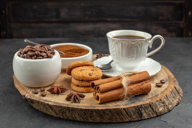 Vista inferior de la taza de café, anís, granos de café tostados y cacao en tazones, palitos de canela, galletas sobre tablero de madera en la oscuridad.