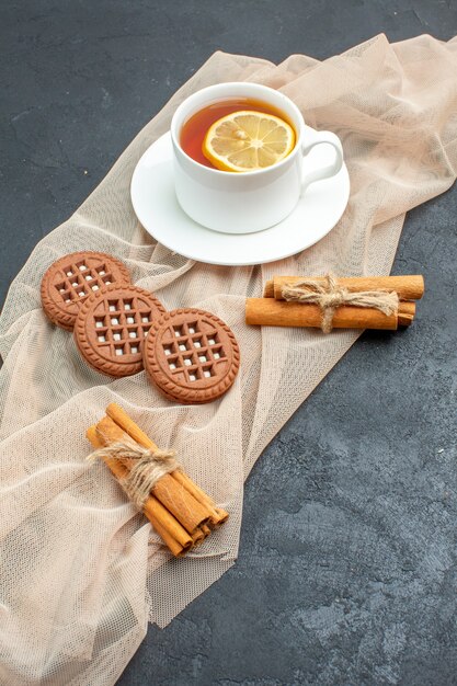 Vista inferior de una taza de té con limón canela palitos de galletas en un chal beige sobre una superficie oscura