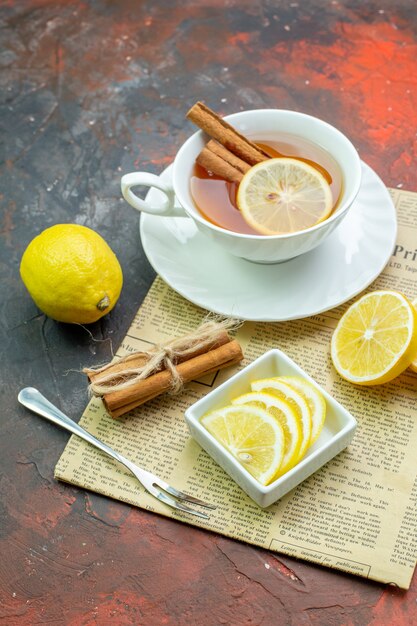 Vista inferior de la taza de té con rodajas de limón canela en un tazón pequeño tenedor palitos de canela atados con una cuerda en el periódico en la mesa de color rojo oscuro