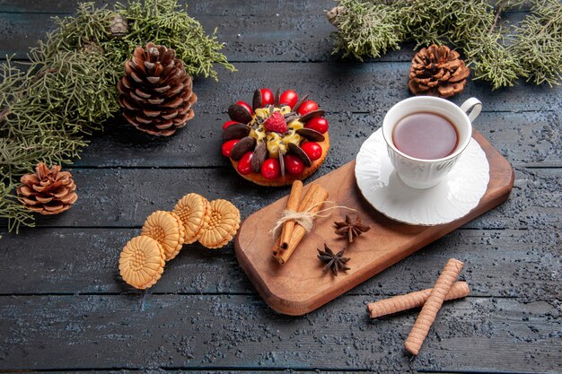 Vista inferior de una taza de té de semillas de anís y canela en un plato de madera para servir piñas pastel de bayas y diferentes galletas sobre fondo oscuro