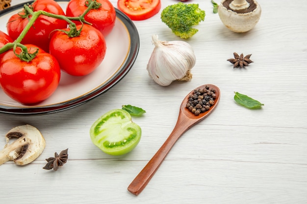 Vista inferior de tomates rojos en un plato blanco, setas de ajo, anís, pimientos negros en una cuchara de madera sobre la mesa gris