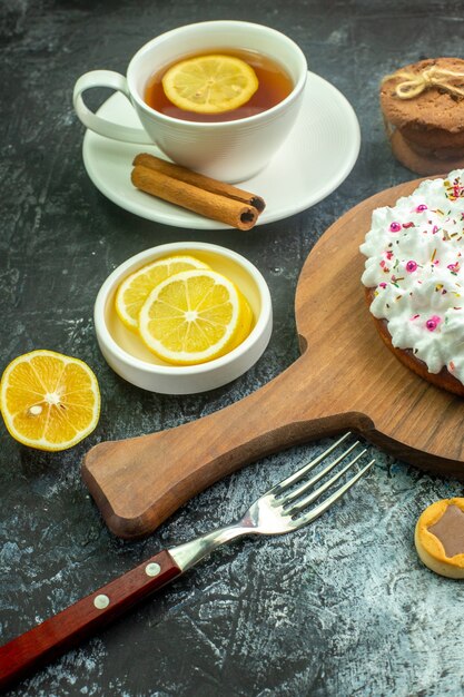 Vista inferior de la torta con crema pastelera en un tablero de madera para servir galletas tenedor palitos de canela taza de té con sabor a limón y canela en la mesa gris