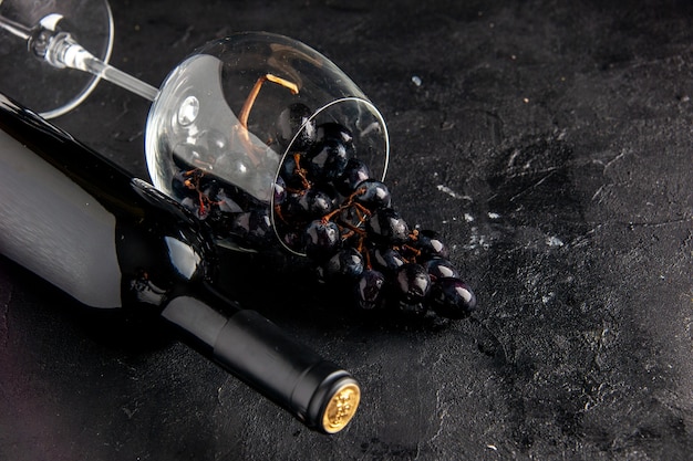 Vista inferior de uvas negras en copa de vino volcada botella de vino sobre mesa oscura