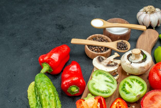 Vista inferior verduras champiñones tomates pimientos rojos sobre tabla de cortar ajo pimienta negra y sal en tazones cucharas de madera pepinos en la mesa oscura espacio libre