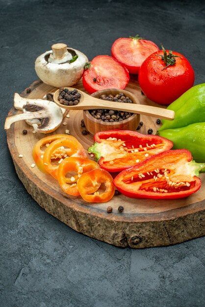 Vista inferior de verduras frescas, setas, pimienta negra en un tazón, cuchara de madera, tomates rojos, pimientos sobre tablero de madera en la mesa oscura.