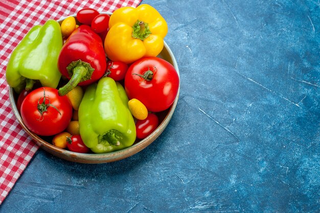 Vista inferior verduras frescas tomates cherry diferentes colores pimientos tomates cumcuat en placa sobre mantel a cuadros rojo blanco sobre mesa azul