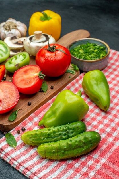 Vista inferior de verduras tomates pimientos en tabla de cortar verduras en un tazón pepinos sobre un mantel rojo sobre una mesa negra