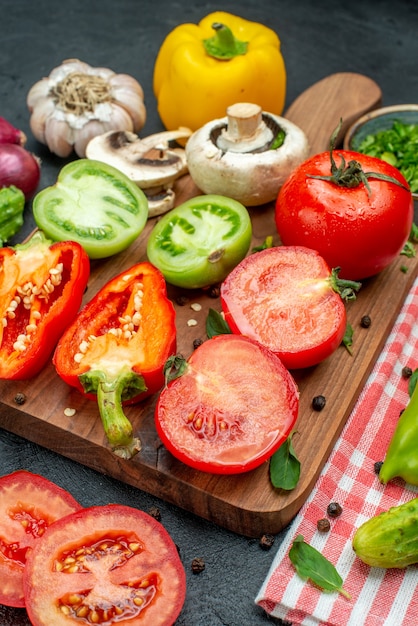 Vista inferior de verduras tomates verdes y rojos pimiento amarillo en la tabla de cortar verduras en un tazón cuchillo pepinos sobre un mantel rojo sobre una mesa negra