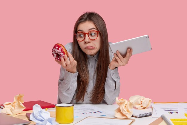 La vista interior de una mujer vacilante sostiene el panel táctil en una mano, una deliciosa rosquilla en la otra, se ve confusa, duda si come comida chatarra, se rompe después del papeleo