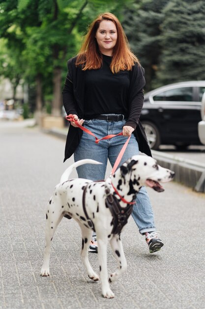 Vista de la joven mujer caucásica caminando en la ciudad durante la mañana con perro dálmata