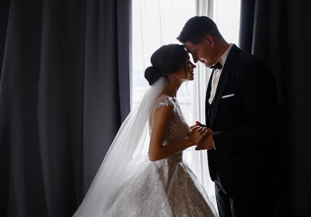 Vista lateral de una adorable pareja vestida de festival de pie cara a cara en la habitación cerrando los ojos tomándose de la mano y sintiéndose feliz durante el día de la boda