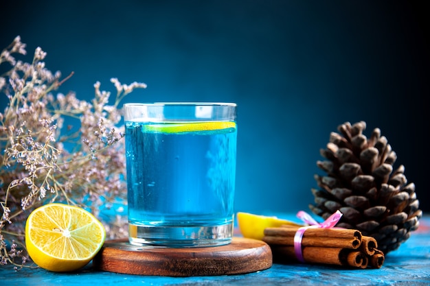 Vista lateral de un agua sin gas con limón en un vaso sobre tabla de cortar de madera y cono de coníferas canela limas sobre fondo azul.