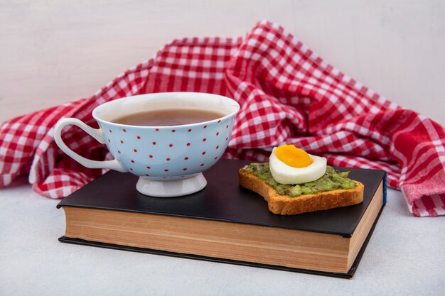 Vista lateral de aguacate en un pan con huevo escalfado y una taza de té sobre el libro sobre mantel rojo y superficie blanca