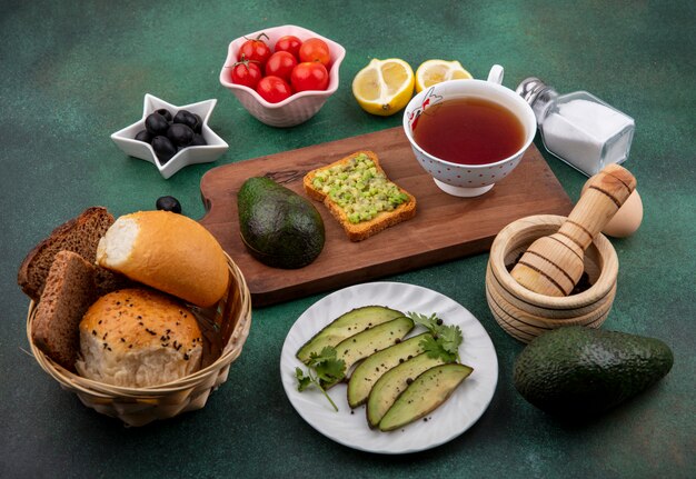 Vista lateral de aguacate sobre una tabla de cocina de madera con una taza de té con aceitunas negras, tomates, limones, un cubo de panes en la superficie verde