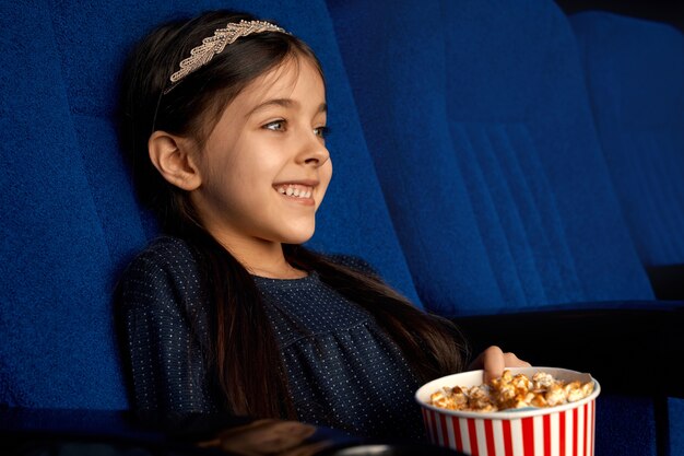 Vista lateral de la alegre chica morena con cola de caballo riéndose de comedia divertida en el cine. Niña feliz comiendo palomitas de maíz y relajándose el fin de semana