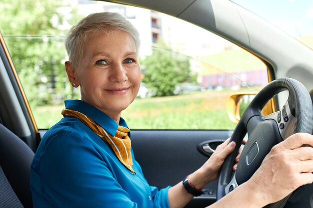 Vista lateral de la alegre mujer de mediana edad dentro del coche en el asiento del conductor con las manos en el volante