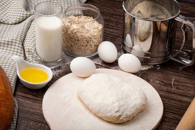 Vista lateral de alimentos como mantequilla derretida leche huevo con copos de avena y masa espolvoreada con harina en la tabla de cortar sobre fondo de madera