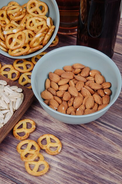 Vista lateral de almendras en un tazón y mini pretzels con una botella de cerveza en rústico