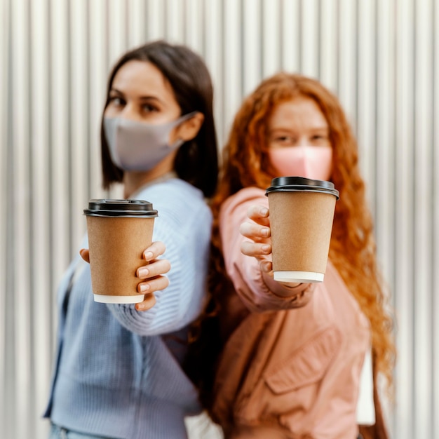 Vista lateral de amigas desenfocadas con máscaras faciales al aire libre sosteniendo tazas de café