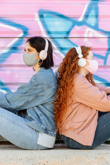 Vista lateral de amigas con máscaras faciales al aire libre escuchando música con auriculares
