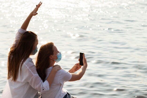 Vista lateral de amigas con máscaras faciales tomando selfie junto al lago