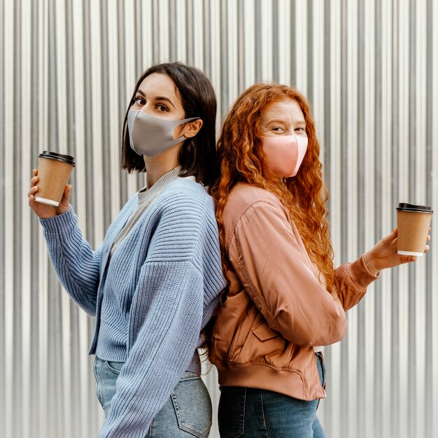 Vista lateral de amigas con mascarillas al aire libre sosteniendo tazas de café