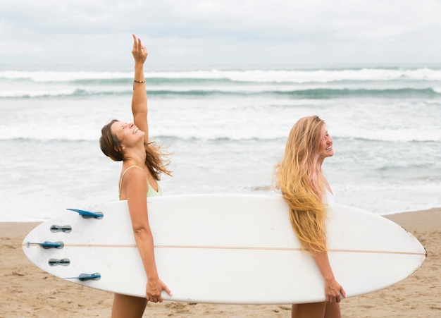 Vista lateral de amigas sosteniendo una tabla de surf en la playa.