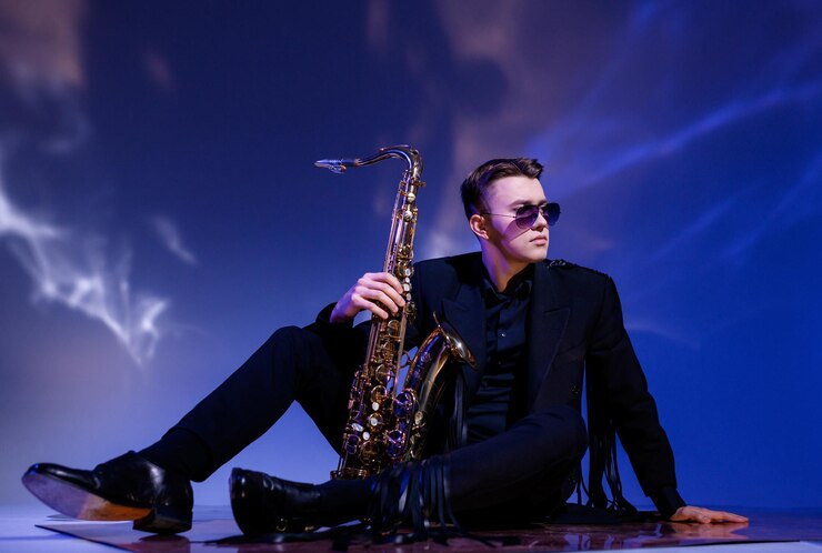 Foto gratuito vista lateral de un apuesto músico masculino vestido con un moderno traje negro y gafas de sol sentado en el suelo y sosteniendo un saxofón dorado mientras mira seriamente hacia otro lado y posa
