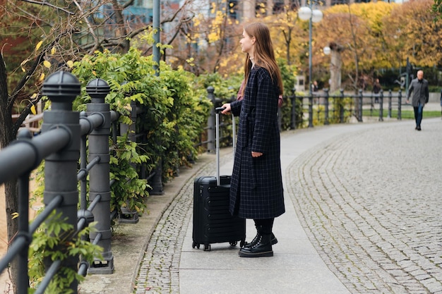 Vista lateral de una atractiva chica casual con abrigo caminando cuidadosamente por la calle con una pequeña maleta negra