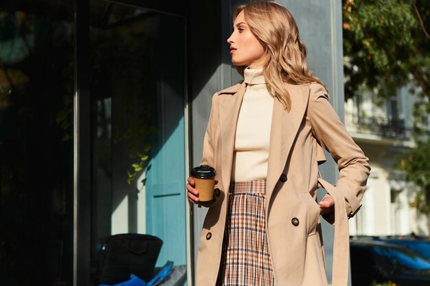 Vista lateral de una atractiva chica rubia elegante con abrigo beige con café para caminar por la calle de la ciudad