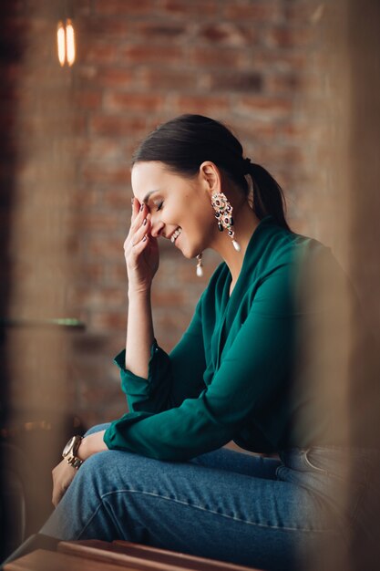 Vista lateral de la atractiva mujer de cabello oscuro en hermosos aretes, blusa esmeralda y jeans con reloj de moda riendo con la mano en la cara. Ella está sentada en un banco de madera contra la pared de ladrillo en el interior.